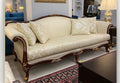 An ornate three-seat sofa with cream damask upholstery and carved dark wood legs, shown in a showroom setting.