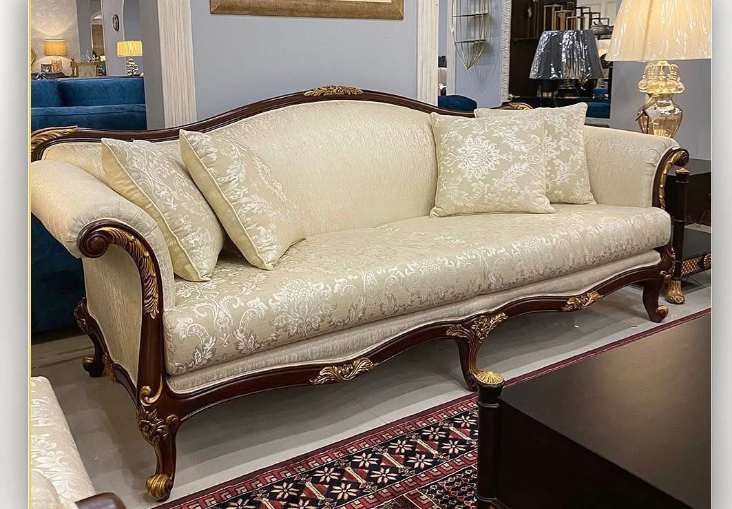 An ornate three-seat sofa with cream damask upholstery and carved dark wood legs, shown in a showroom setting.