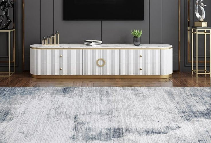 A modern white TV console with gold accents and a marble-look top, styled in a contemporary living room setting with a blue-gray rug, dark gray wall, and wooden floor.
