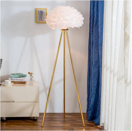 A modern tripod floor lamp with a fluffy white cloud-shaped shade and slim gold metal legs, shown in a styled room setting with blue and white curtains, a wooden floor, and a cream side table.