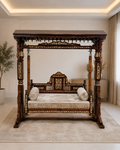 An ornate dark wood four-poster canopy daybed with intricate gold-toned carvings, upholstered with a cream velvet cushion and two matching cylindrical pillows, set in a bright, modern room on a beige rug.