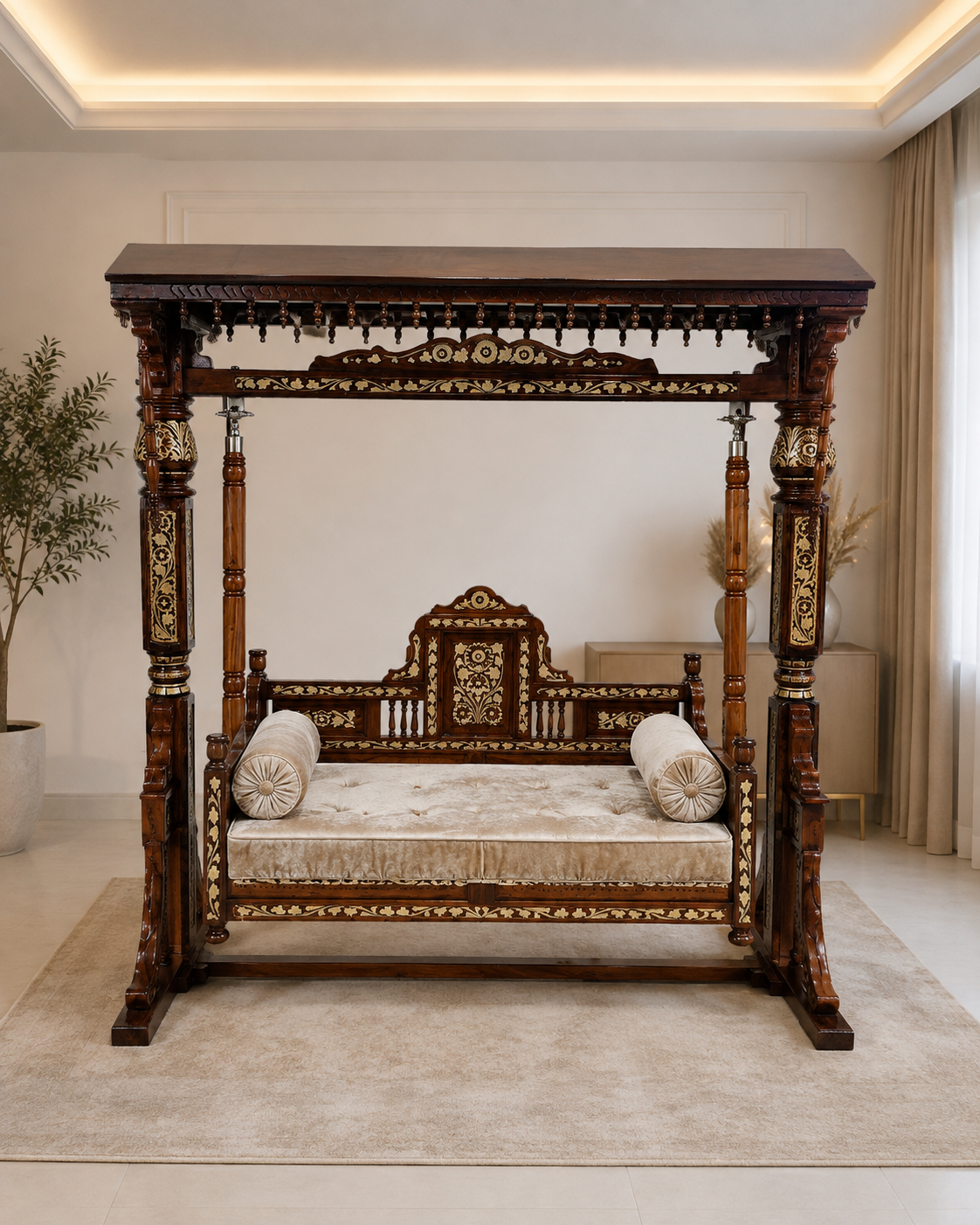 An ornate dark wood four-poster canopy daybed with intricate gold-toned carvings, upholstered with a cream velvet cushion and two matching cylindrical pillows, set in a bright, modern room on a beige rug.