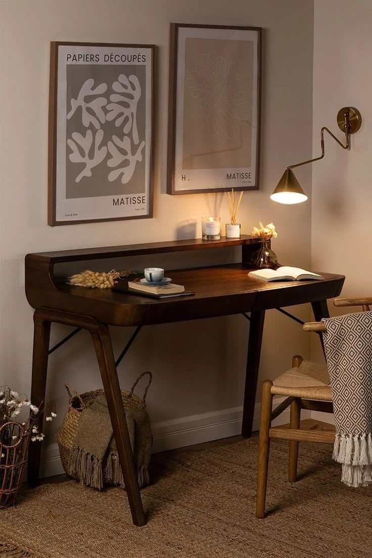 A dark wood writing desk styled in a cozy home office setting with a wooden chair, a brass wall lamp, and two framed Matisse art prints on the wall.