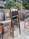 An antique-style gold-framed full-length standing mirror with an ornate carved top, photographed in a workshop or antique store setting with blurred furniture and greenery in the background.