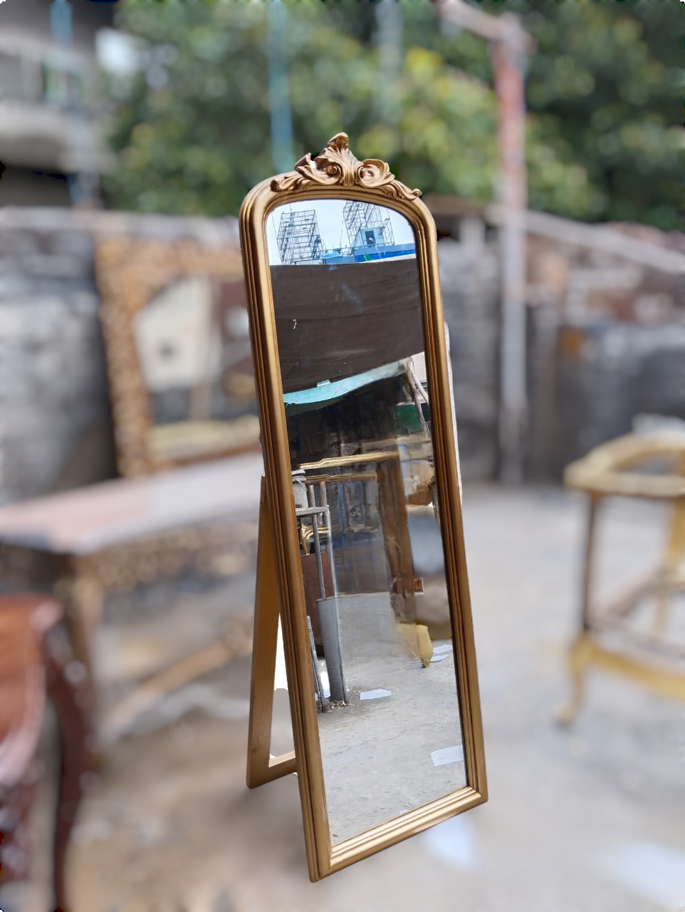 An antique-style gold-framed full-length standing mirror with an ornate carved top, photographed in a workshop or antique store setting with blurred furniture and greenery in the background.