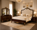 An ornate classic bedroom set featuring a dark wood bed with a tufted cream headboard and gold accents, a matching wooden dresser with a large mirror, bedside tables with lamps, and a beige patterned rug in a luxury decorated room.