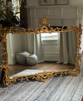 An ornate gold-framed floor mirror with intricate baroque-style scrollwork, leaning against a white fireplace in a styled room with wood floors and a patterned rug.