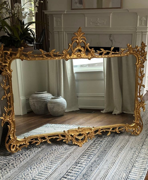 An ornate gold-framed floor mirror with intricate baroque-style scrollwork, leaning against a white fireplace in a styled room with wood floors and a patterned rug.