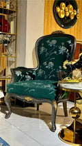 An ornate high-back accent chair with dark green floral velvet upholstery and carved dark wood legs, styled in a luxury interior next to a gold side table.