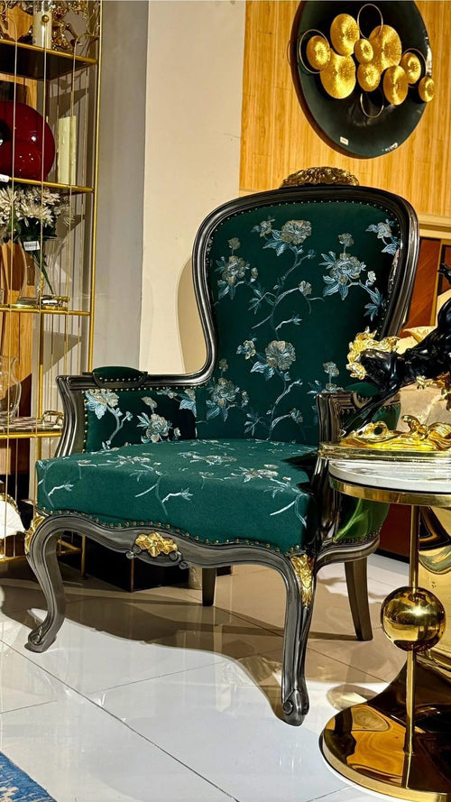 An ornate high-back accent chair with dark green floral velvet upholstery and carved dark wood legs, styled in a luxury interior next to a gold side table.